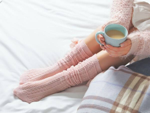Woman relaxing at cozy home atmosphere on the bed. Young woman with cup of cocoa or coffee in hands and cookies enjoying comfort. Soft light and comfy lifestyle concept.