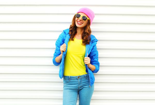 Fashion pretty smiling woman model in colorful clothes posing over white background wearing a pink hat yellow sunglasses and blue jacket