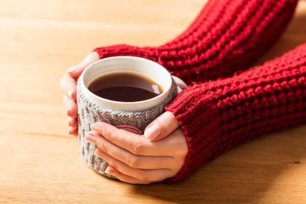 Hot mug of tea warming woman's hands in retro woollen jumper. Wooden table