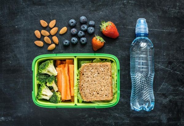 School lunch box with sandwich, vegetables, water, almonds and fruits on black chalkboard background. Healthy eating habits concept. Flat lay composition (from above, top view).